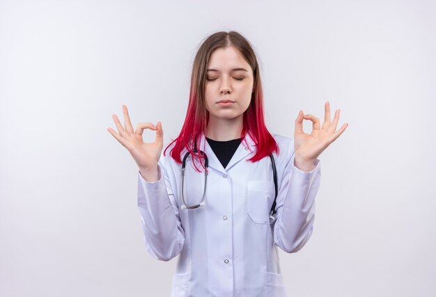 Con gli occhi chiusi giovane ragazza medico indossando stetoscopio abito medico che mostra il gesto di pace con entrambe le mani sul muro bianco isolato