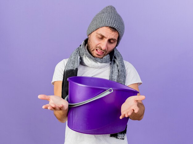 Con gli occhi chiusi giovane uomo malato che indossa un cappello invernale con sciarpa tenendo il secchio di plastica e tendendo le mani alla telecamera isolata su sfondo viola