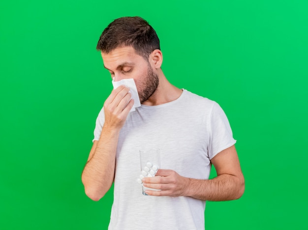 Con gli occhi chiusi giovane uomo malato pulendosi il naso con il tovagliolo e tenendo le pillole con un bicchiere d'acqua isolato su sfondo verde