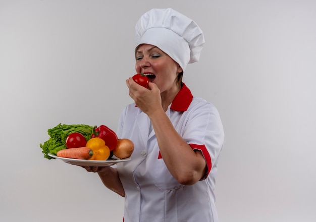 Con gli occhi chiusi il cuoco femminile di mezza età in uniforme del cuoco unico che tiene le verdure sul piatto morde il pomodoro con lo spazio della copia