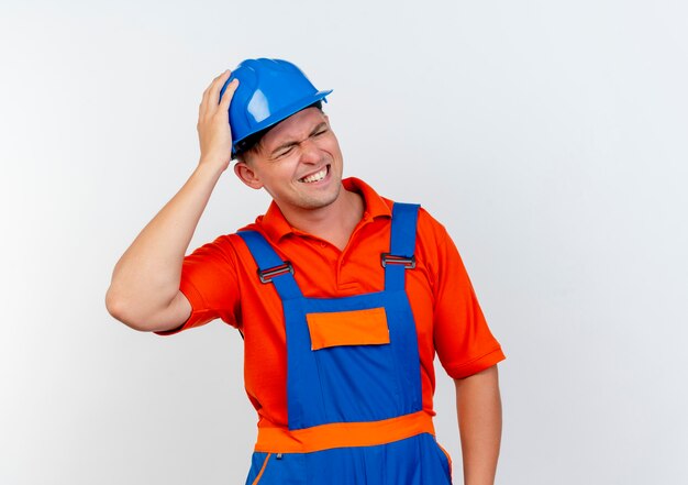 Con gli occhi chiusi, il giovane costruttore maschio interessato indossa l'uniforme e il casco di sicurezza mettendo la mano sulla testa su bianco