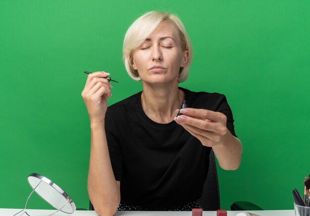 Con gli occhi chiusi la giovane bella ragazza si siede al tavolo con gli strumenti di trucco che porge il mascara alla macchina fotografica isolata su fondo verde