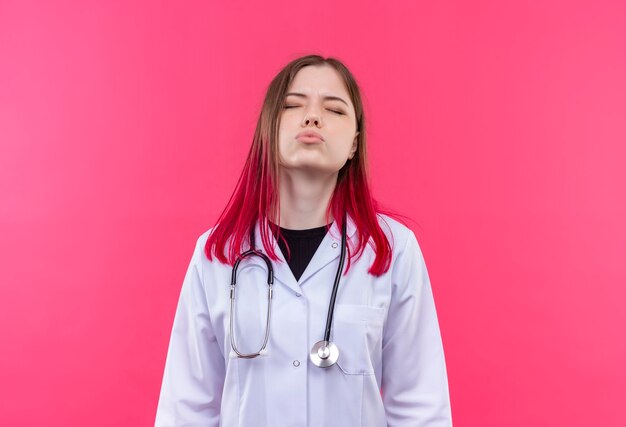 Con gli occhi chiusi la giovane ragazza del medico che indossa l'abito medico dello stetoscopio che mostra il gesto di bacio sulla parete isolata rosa