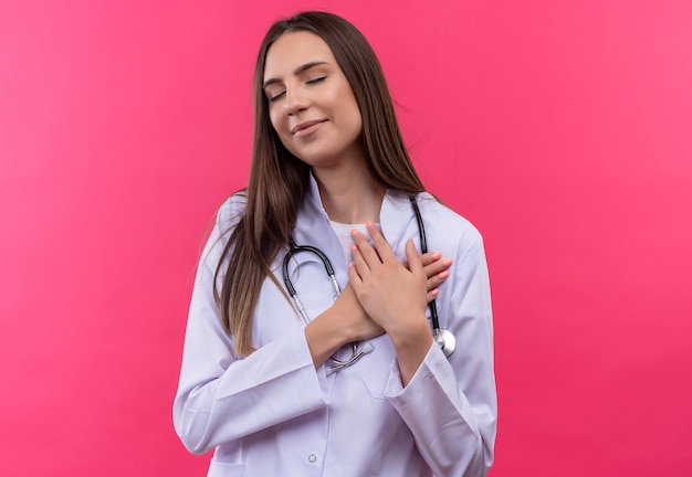 Con gli occhi chiusi la ragazza giovane medico soddisfatta che indossa l'abito medico dello stetoscopio mise le mani sul cuore sul muro rosa isolato