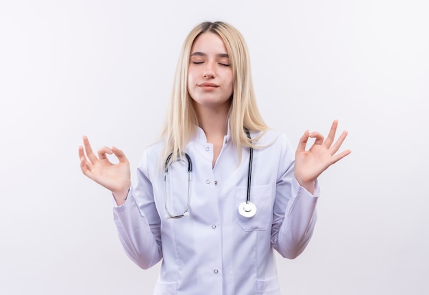 Con gli occhi chiusi medico giovane ragazza bionda che indossa lo stetoscopio e abito medico che mostra okey gesto con entrambe le mani sul muro bianco isolato