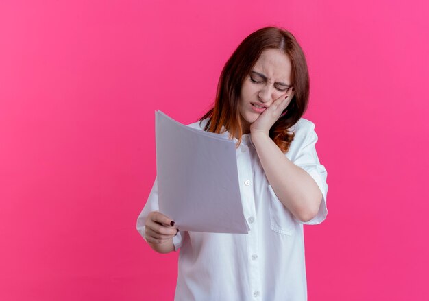 Con gli occhi chiusi ragazza giovane rossa stanca che tiene la carta e che mette la mano sulla guancia isolata sul colore rosa con lo spazio della copia