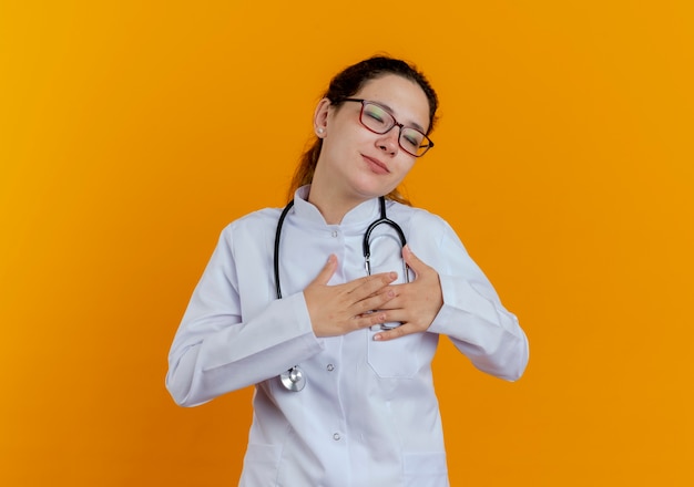 Con gli occhi chiusi soddisfatto giovane medico femminile che indossa abito medico e stetoscopio con gli occhiali che mettono le mani sul cuore isolato