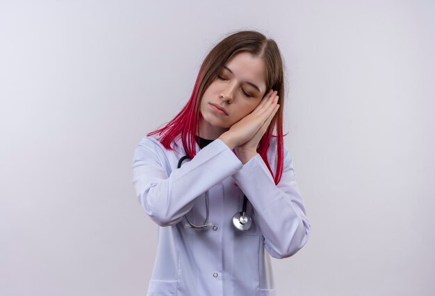 Con la veste medica da portare dello stetoscopio della giovane ragazza degli occhi chiusi che mostra il gesto di sonno sulla parete bianca isolata