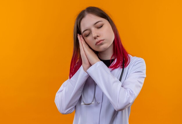 Con la veste medica da portare dello stetoscopio della giovane ragazza di medico degli occhi chiusi che mostra il gesto di sonno sulla parete arancione isolata