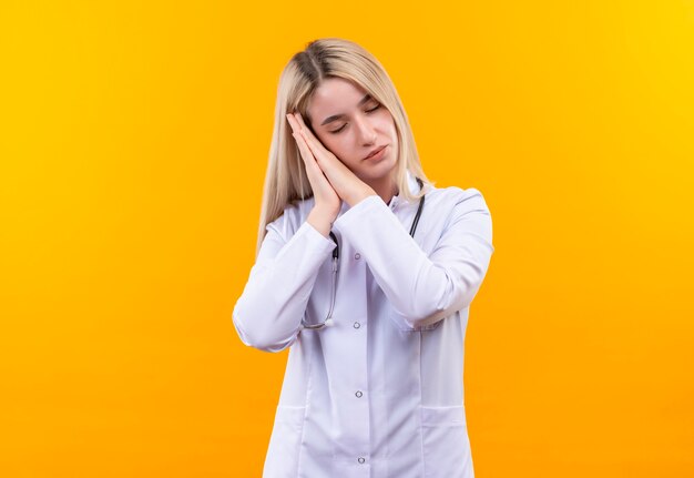 Con lo stetoscopio d'uso della ragazza del medico degli occhi chiusi in abito medico che mostra il gesto di sonno sulla parete gialla isolata