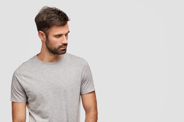 Concentrato bel giovane con la barba lunga messo da parte, ha un corpo muscoloso, vestito con una maglietta grigia casual