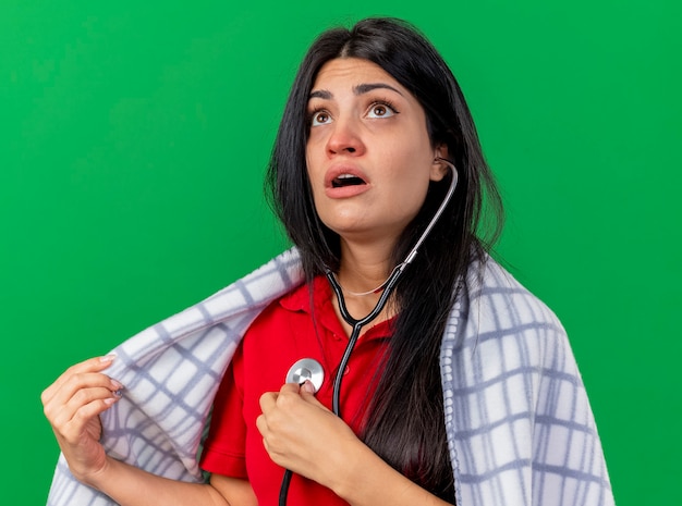 Concentrato di giovani caucasici ragazza malata che indossa uno stetoscopio avvolto in plaid ascoltando il suo battito cardiaco cercando afferrando plaid isolato su sfondo verde
