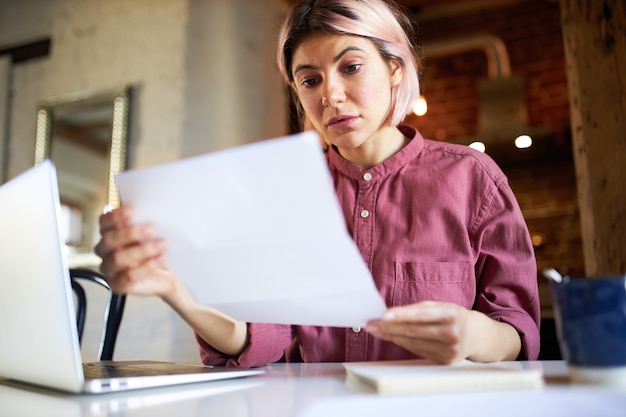 Concentrato di successo giovane economista femminile analizzando dati importanti, seduto davanti al computer portatile aperto, studiando il documento contabile.