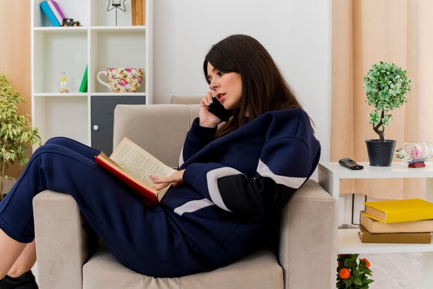 Concentrato giovane donna abbastanza caucasica che si siede sulla poltrona nel soggiorno progettato parlando al telefono con il libro sulle gambe guardando il libro e toccandolo