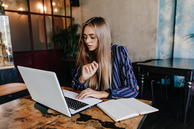 Concentrato giovane donna che lavora nella caffetteria preferita. Tiro al coperto di affascinante studentessa seria utilizzando laptop.