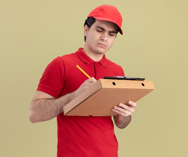 Concentrato giovane fattorino caucasico in uniforme blu e cappuccio che tiene il pacchetto della pizza e appunti su di esso scrivendo con la matita negli appunti