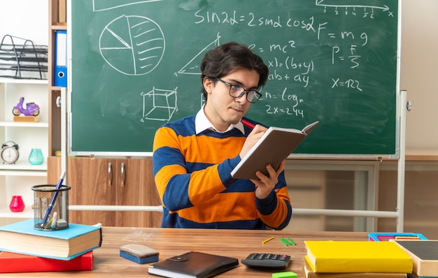 Concentrato giovane insegnante di geometria con gli occhiali seduto alla scrivania con materiale scolastico in classe scrivendo su un blocco note con penna