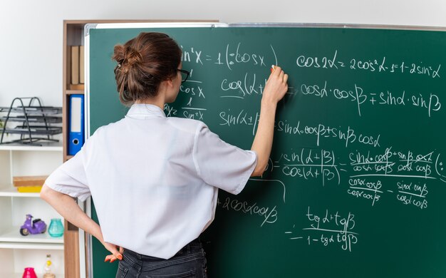 Concentrato giovane insegnante di matematica femminile bionda con gli occhiali in piedi dietro la vista davanti alla lavagna che tiene il gesso tenendo la mano sulla vita risolvendo il problema di matematica in classe