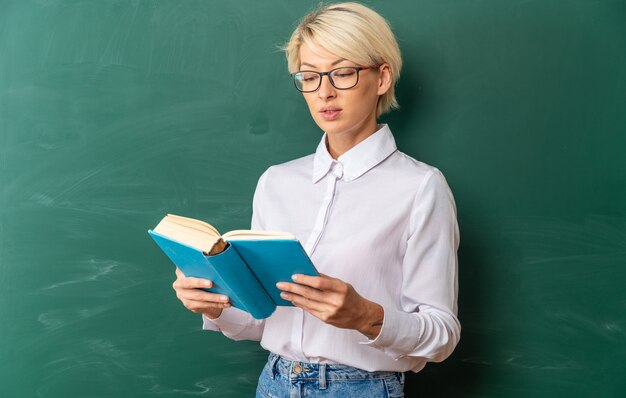 Concentrato giovane insegnante di sesso femminile bionda con gli occhiali in aula in piedi di fronte alla lavagna che tiene e legge un libro