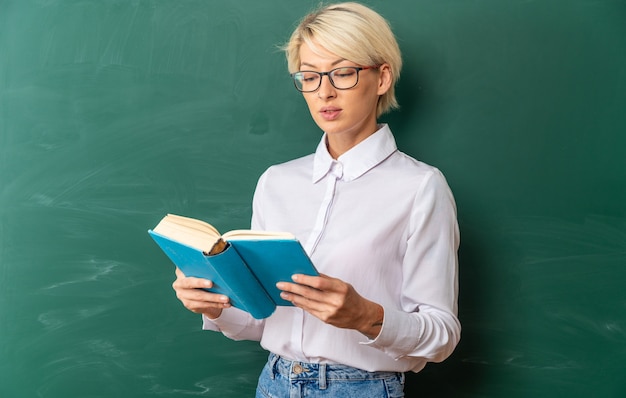 Concentrato giovane insegnante di sesso femminile bionda con gli occhiali in aula in piedi di fronte alla lavagna che tiene e legge un libro