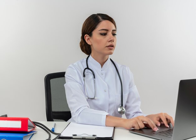 Concentrato giovane medico femminile che indossa abito medico e stetoscopio seduto alla scrivania con strumenti medici utilizzando laptop isolato sul muro bianco