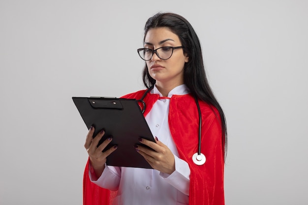 Concentrato giovane ragazza caucasica del supereroe che indossa lo stetoscopio che tiene e che esamina appunti isolato su priorità bassa bianca con lo spazio della copia