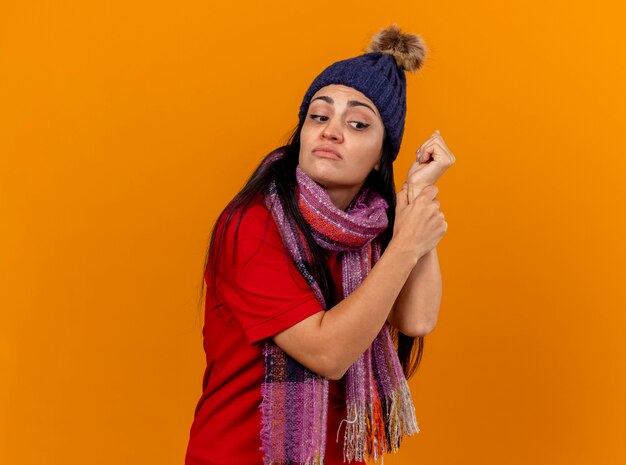 Concentrato giovane ragazza malata caucasica che indossa cappello invernale e sciarpa controllo impulso guardando lato isolato sulla parete arancione con lo spazio della copia