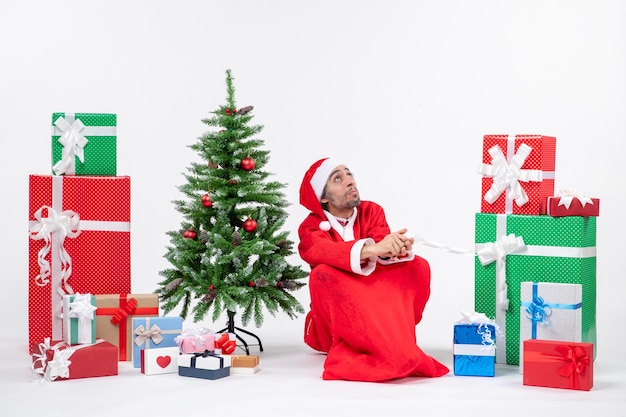 Concentrato giovane uomo vestito da Babbo Natale con doni e albero di Natale decorato seduto per terra su sfondo bianco