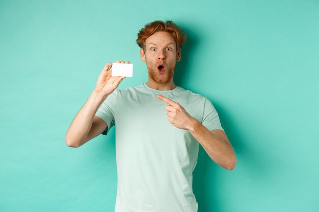 Concetto di acquisto. Bell'uomo dai capelli rossi in maglietta che mostra una carta di credito in plastica e sorride, in piedi su sfondo turchese