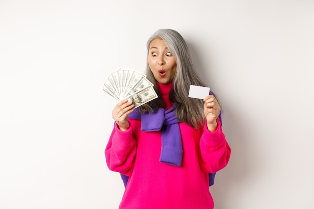 Concetto di acquisto. Donna anziana asiatica fortunata che guarda stupita i soldi e mostra una carta di credito in plastica, in piedi su uno sfondo bianco.