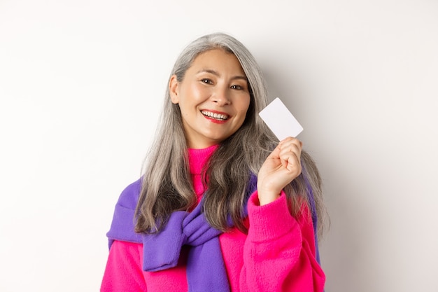 Concetto di acquisto. Elegante donna anziana asiatica che sorride e mostra una carta di credito in plastica, pagando senza contatto, in piedi su sfondo bianco.