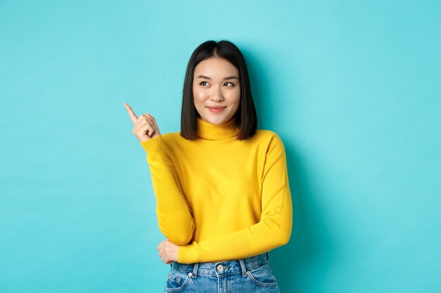 Concetto di acquisto. Elegante modello femminile asiatico in maglione giallo, sorridente e puntando il dito a sinistra, mostrando pubblicità con faccia soddisfatta, in piedi su sfondo blu