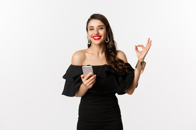 Concetto di acquisto online. donna in abito nero alla moda, trucco, che mostra l'approvazione del segno ok e utilizza l'app del telefono cellulare, sfondo bianco.
