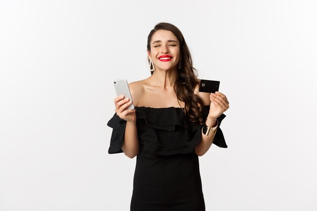 Concetto di acquisto online. Moda donna in abito nero, tenendo la carta di credito con lo smartphone, guardando soddisfatta, in piedi su sfondo bianco.