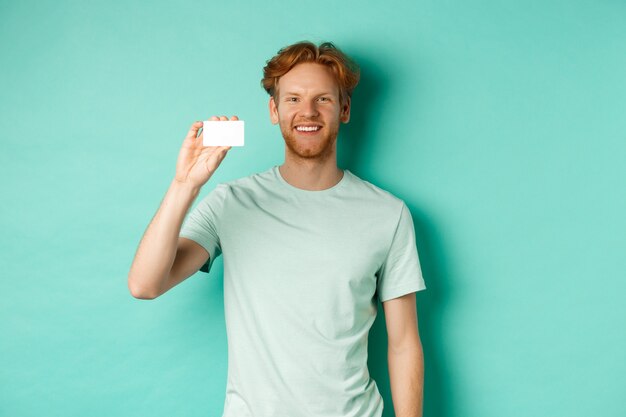 Concetto di acquisto. Uomo bello rosso in t-shirt che mostra carta di credito in plastica e sorridente, in piedi su sfondo turchese.