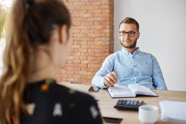 Concetto di affari. Colloquio di lavoro. Responsabile senior maschio barbuto adulto in vetri e camicia che si siedono nell'ufficio leggero davanti alla donna caucasica castana, facente domande sul posto di lavoro precedente