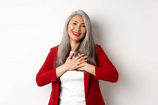 Concetto di affari. Donna matura asiatica con labbra rosse e giacca sportiva, mano nella mano sul cuore e sorridente grato, guardando grato alla telecamera, in piedi su sfondo bianco.