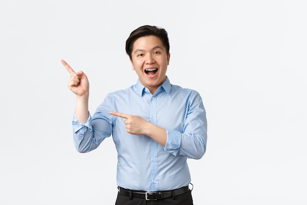 Concetto di affari, finanza e persone. Venditore asiatico allegro in camicia blu con bretelle per denti, puntando le dita nell'angolo in alto a sinistra e sorridendo eccitato, mostrando l'annuncio, consigliando il prodotto.