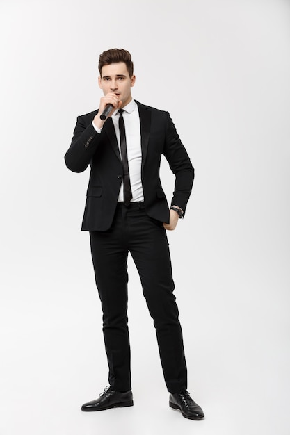 Concetto di affari: Ritratto a figura intera giovane in abito nero tiene in mano un microfono, canta e posa su uno sfondo bianco