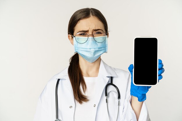 Concetto di assistenza medica online triste e cupo giovane medico in maschera facciale che mostra lo schermo dello smartphone con ...
