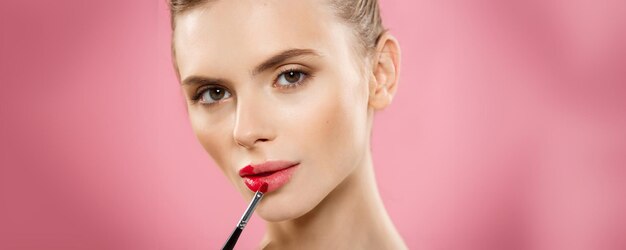 Concetto di bellezza Donna che applica rossetto rosso con sfondo rosa per studio Bella ragazza fa il trucco