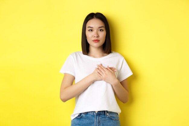 Concetto di bellezza e moda. Bella donna asiatica che si tiene per mano sul cuore e guarda premurosa la macchina fotografica, conservando i ricordi nell'anima, in piedi su sfondo giallo