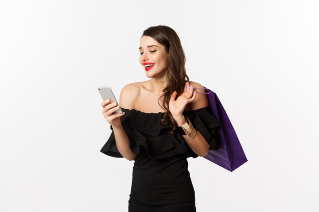 Concetto di bellezza e shopping. Splendida donna in elegante abito nero e trucco, ordina online su smartphone, tiene in mano la borsa e sorride, in piedi su sfondo bianco.