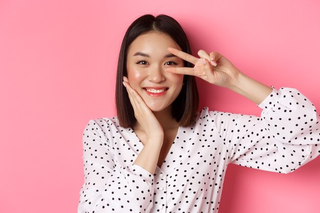 Concetto di bellezza e stile di vita. Primo piano di una donna asiatica carina che mostra segno di pace e tocca la guancia, sorride felice alla telecamera, in piedi su sfondo rosa