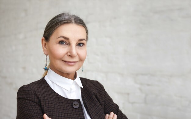 Concetto di bellezza, stile, moda e età. Close up ritratto di positivo elegante femmina di 60 anni con i capelli grigi e la faccia rugosa in posa contro il muro di mattoni bianchi