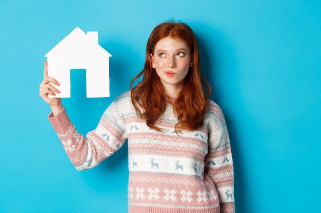 Concetto di bene immobile. Immagine della ragazza rossa premurosa che mostra il modello della casa di carta e pensa, cercando casa o appartamento, in piedi su sfondo blu.
