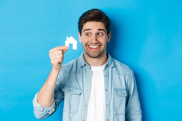 Concetto di bene immobile. Ragazzo eccitato che guarda il modello di una piccola casa e sorride, affitta un appartamento, in piedi su sfondo blu.