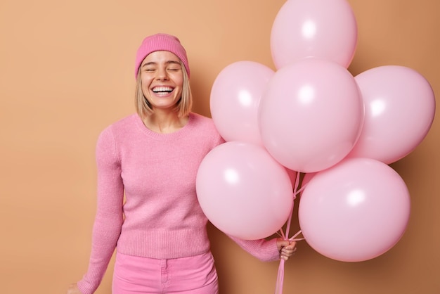 Concetto di celebrazione delle vacanze della gente Una donna dai capelli biondi positiva che fa la doccia per bambini tiene palloncini rosa scopre che avrà una ragazza che indossa abiti casual si sente molto felice isolata sopra il muro beige