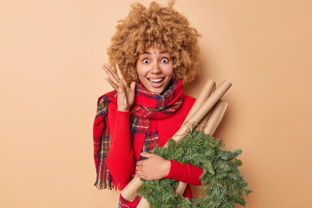 Concetto di celebrazioni per le vacanze di Natale. Eccitato allegro giovane donna alza la mano andando a decorare la ghirlanda di Natale essendo decoratore fiorista indossa sciarpa intorno al collo isolato su sfondo beige.