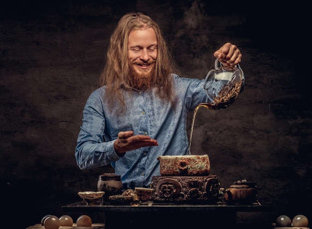 Concetto di cerimonia del tè. Ritratto di un maschio hipster dai capelli rossi felice con i capelli lunghi e la barba piena vestito con una camicia blu, che si diverte a fare il tè, usando un set da tè fatto a mano. Isolato su un dorso strutturato scuro